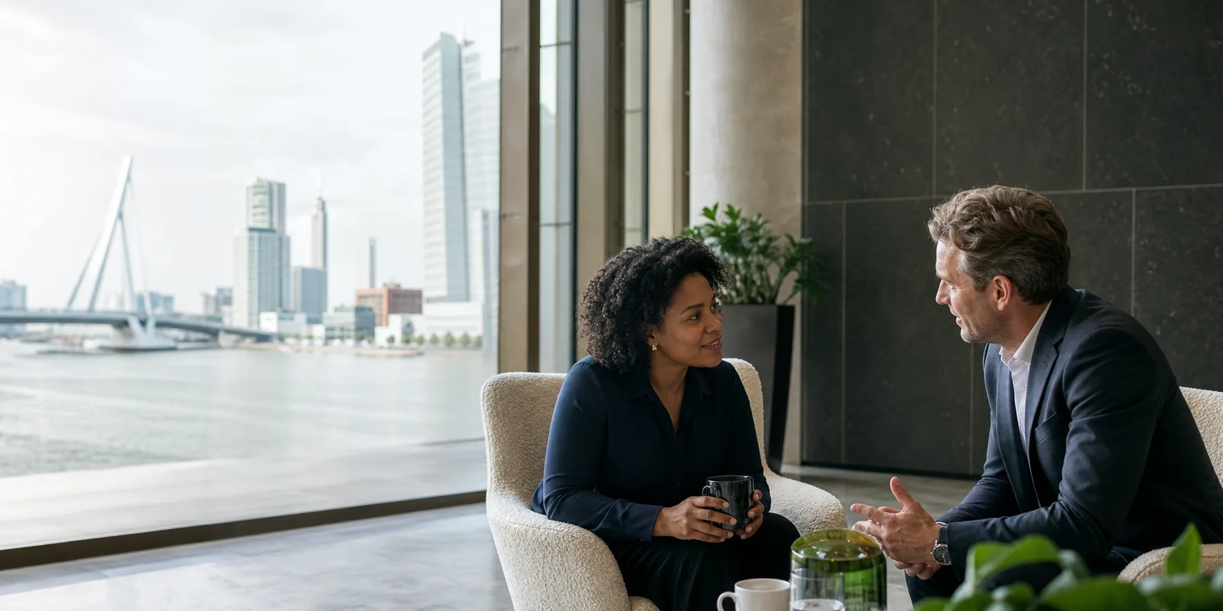 Een consultant en een opdrachtgever in gesprek bij het raam, met de Erasmusbrug op de achtergrond