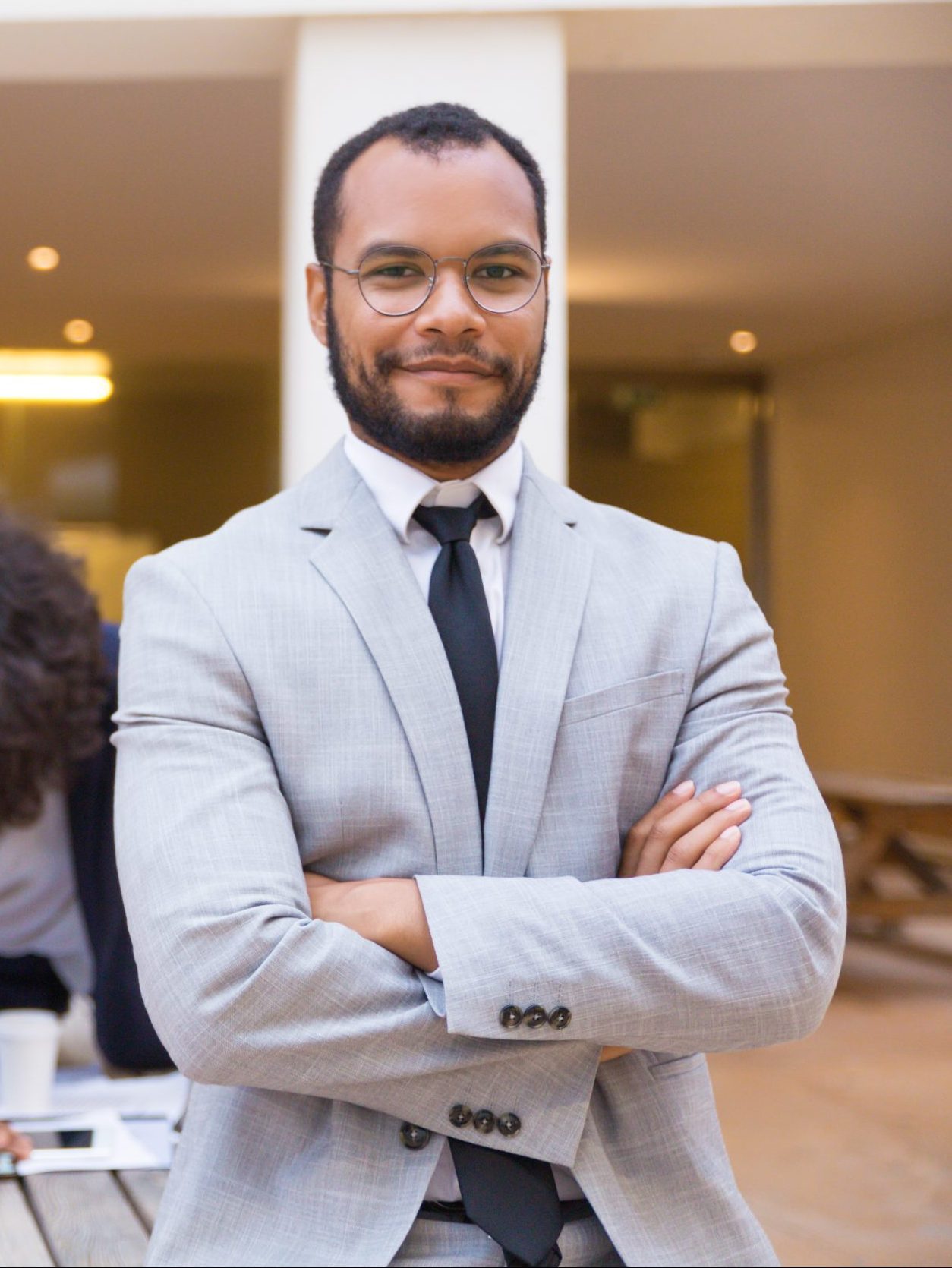 Happy confident male professional posing with arms folded, looking at camera, smiling. Her multiethnic colleagues working in background. Successful team leader concept